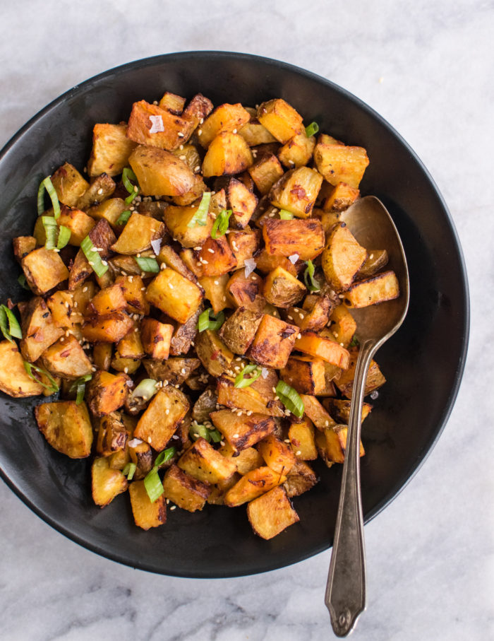 A bowl of spicy roasted potatoes with tahini sauce drizzled on top. Served with scallions and sesame seeds.