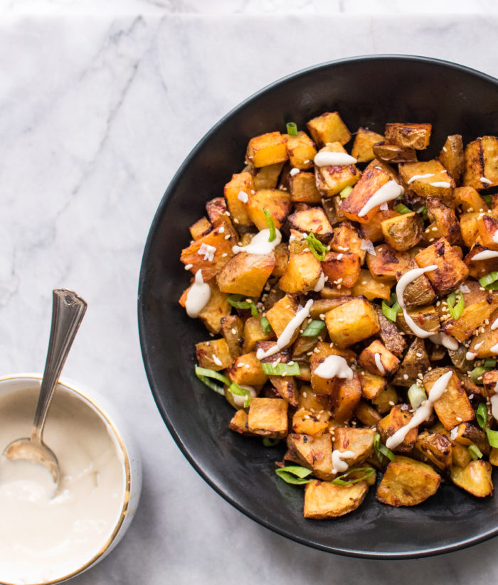 A bowl of spicy roasted potatoes with tahini sauce drizzled on top. Served with scallions and sesame seeds.