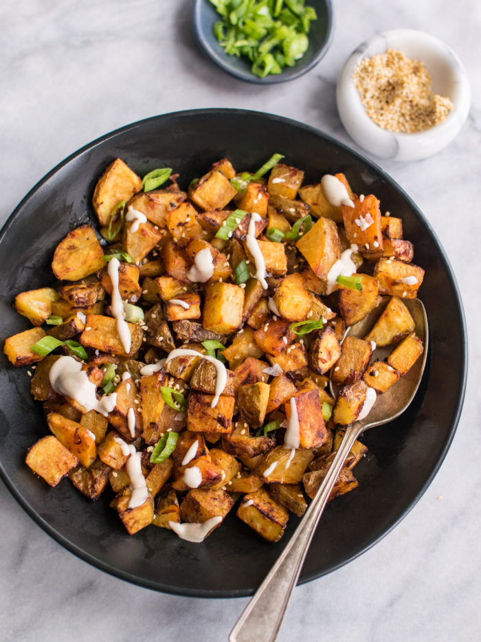 A bowl of spicy roasted potatoes with tahini sauce drizzled on top. Served with scallions and sesame seeds.