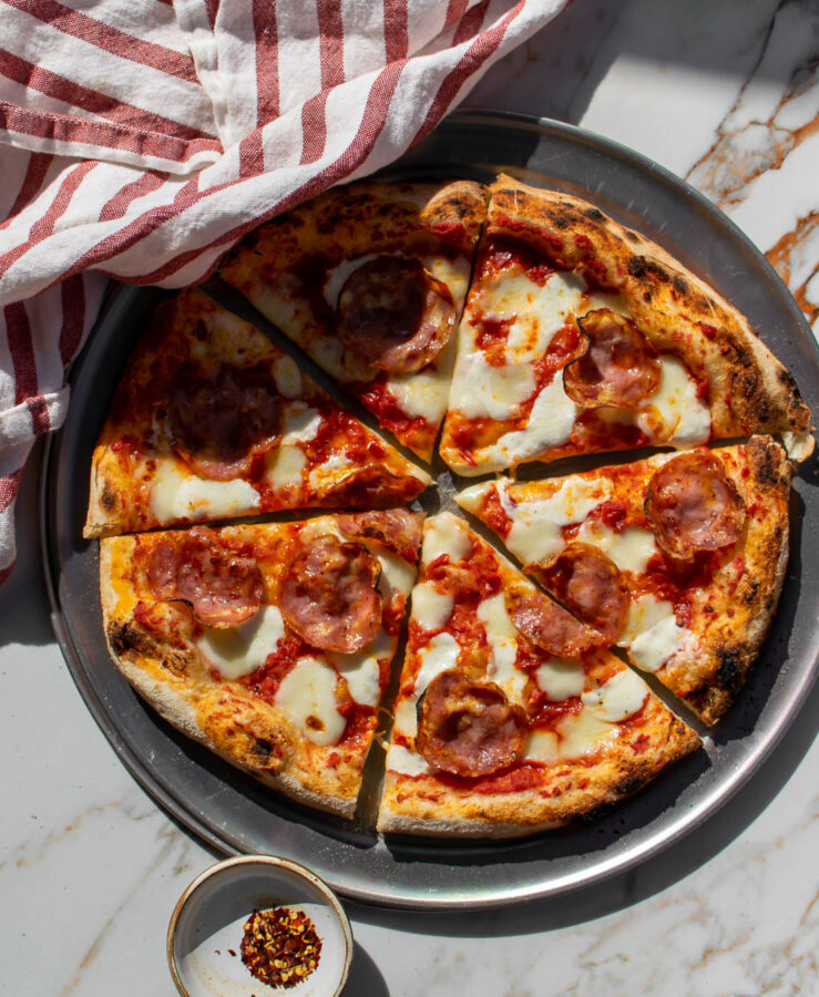 Pizza Diavola with Hot Soppressata sliced on a pizza plate with red pepper flakes in a small bowl to the side.