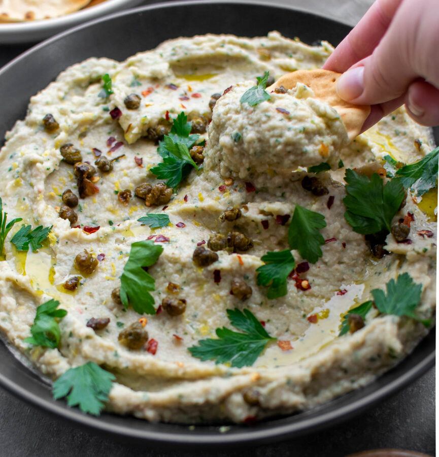 Roasted Garlic and Artichoke White Bean Dip with Fried Capers being eaten with pita chips.